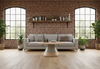 Byorne Coffee Large Table by Quell Design featured in a modern living room with exposed brick walls, wooden flooring, high ceilings, large windows, and lush greenery. The table showcases a rounded oval wood veneer top and bold single pedestal base, combining natural warmth with minimalist Scandinavian design. Crafted from eco-friendly materials, it serves as an elegant focal point that brings balance and sophistication to contemporary interiors.