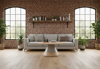 Byorne Coffee Large Table by Quell Design featured in a modern living room with exposed brick walls, wooden flooring, high ceilings, large windows, and lush greenery. The table showcases a rounded oval wood veneer top and bold single pedestal base, combining natural warmth with minimalist Scandinavian design. Crafted from eco-friendly materials, it serves as an elegant focal point that brings balance and sophistication to contemporary interiors.