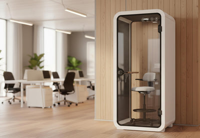 A modern office setting features a pristine white acoustic booth, positioned against a light wooden paneled wall. Inside the booth, the main focal point is a sleek, light grey upholstered ergonomic stool with a small backrest and arm rests, featuring a circular footrest, deisgned for comfort and individual work. In the background, an open-place office is visible with wooden floors, white desks, black office chairs and large windows allowing for natural light.